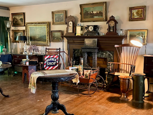 Antique-filled living room featuring a vintage fireplace, classic clocks, framed paintings, wooden furniture, and a retro barber shop sign displayed in a rustic home setting
