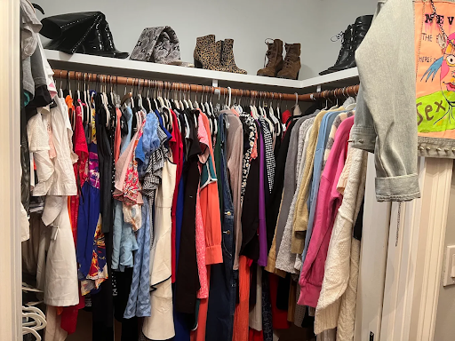 A neatly organized walk-in closet filled with a variety of women’s clothing on hangers, including dresses, jackets, and sweaters in different colors and fabrics. Shelves above hold several pairs of boots and handbags. The space has a lived-in, stylish feel—perfect for showcasing fashion or home organization items for an estate sale listing by Attic to the Basement.